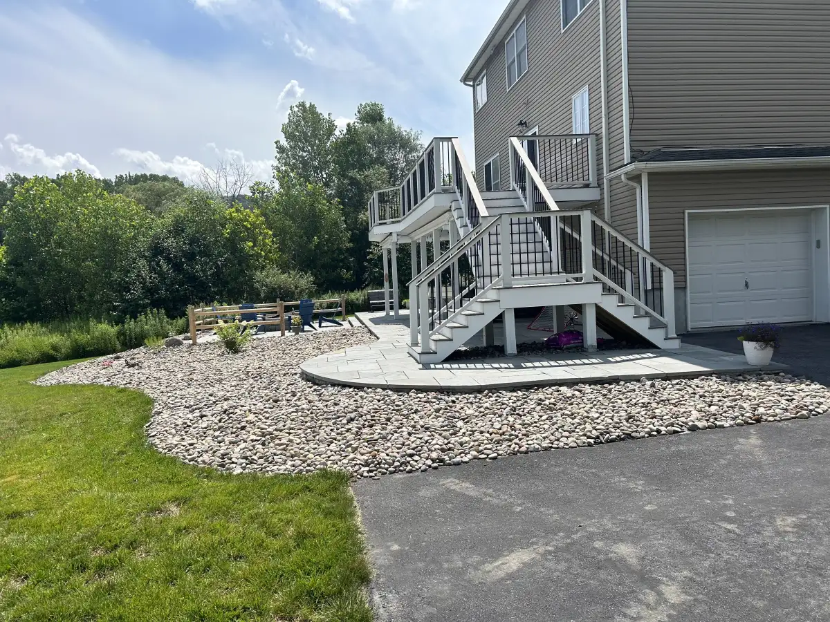 Backyard landscape with river rock, paver patio, and deck stairs by All American Lawn and Landscape